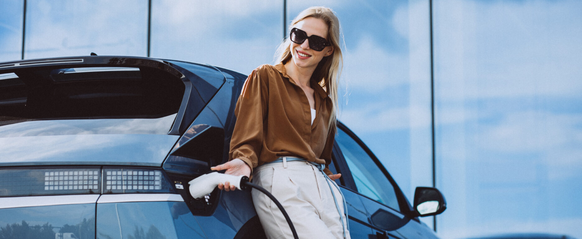 A woman charges an electric car, smiling while holding the charging cable.