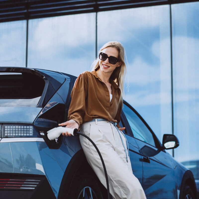 A woman charges an electric car, smiling while holding the charging cable.